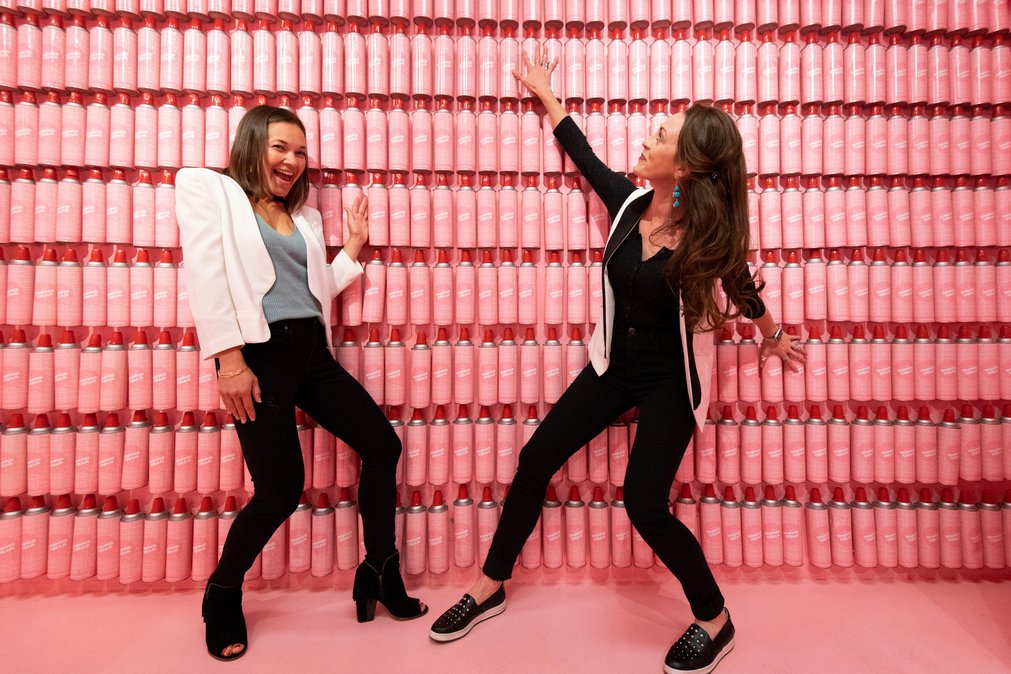 Two women stand by a pink wall of whipped cream cans in the Museum of Ice Cream in San Francisco, California.